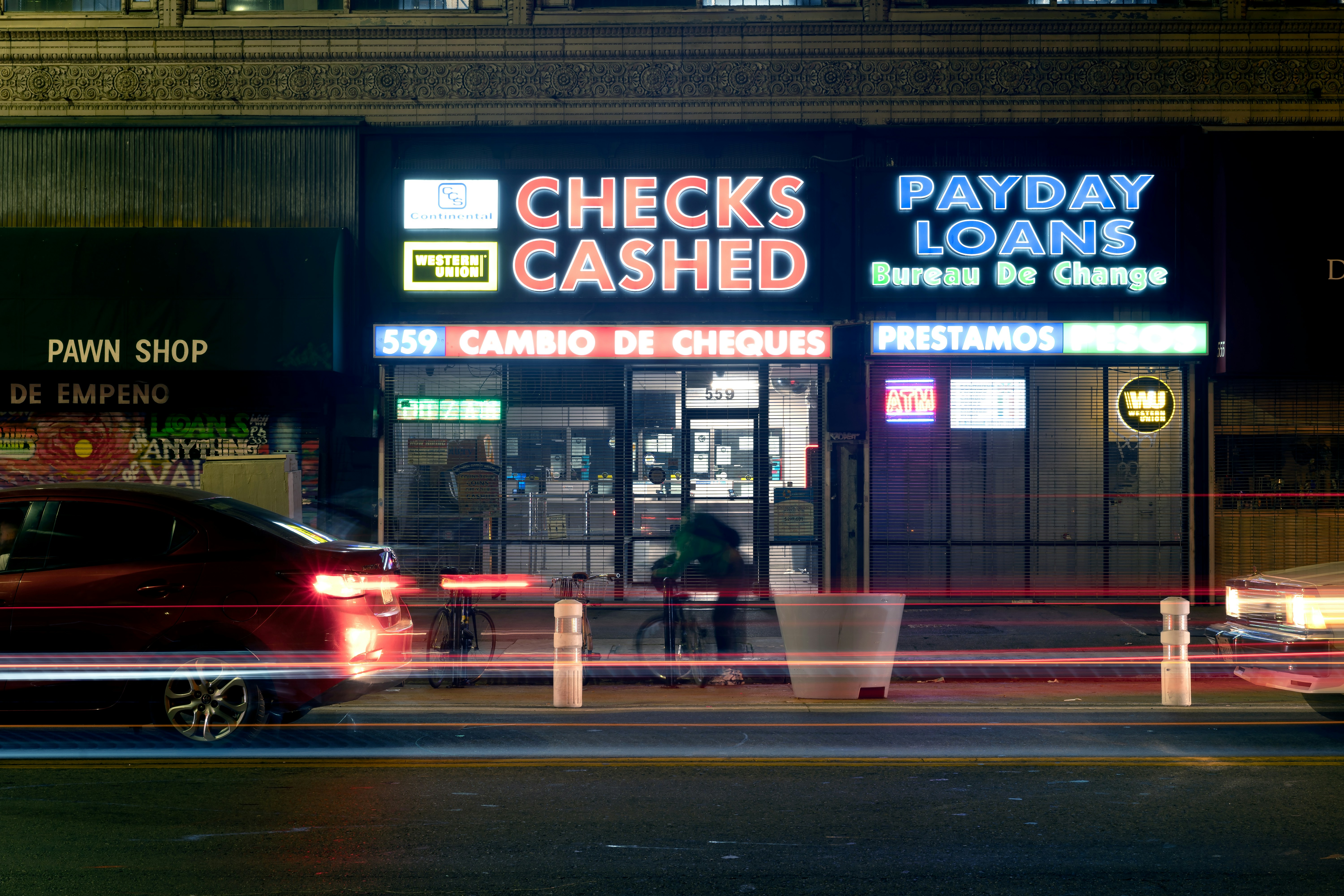 A neon-lit check cashing and payday loan storefront in Downtown Los Angeles with passing traffic and a cyclist blurred in motion. The scene reflects late-night city life and the realities of urban personal finance services in DTLA.